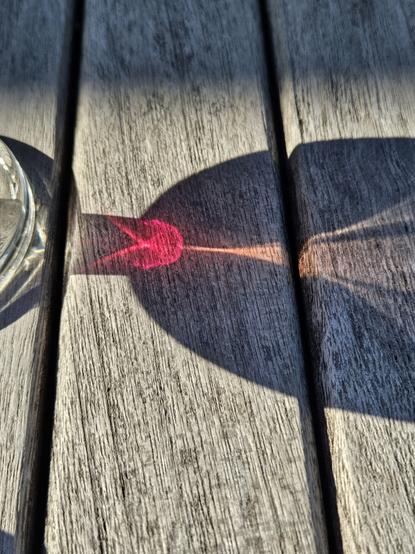 Photo close-up of light patterns cast on an outdoor wooden table by a glass of red wine. First, there's a normal shadow showing a curved wine glass with a short stem. Second, in that shadow at the top of the stem, there's a red mushroom shape (with two angled legs downwards). Third, from the red mushroom upwards in the shadow, there's an expanding curved fan of white light.