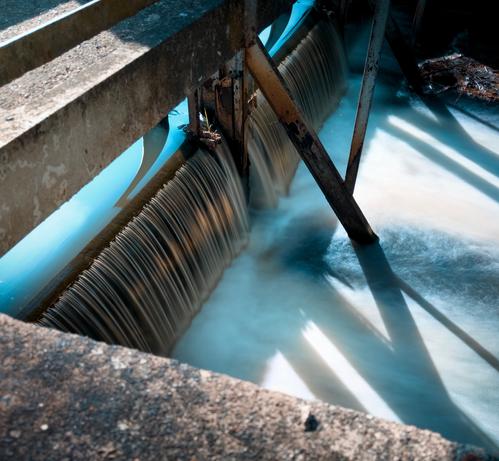 Auf dem Foto ist eine kleine Wasserwehranlage zu sehen. Das Wasser fließt gleichmäßig über eine Kante und bildet einen sanften, seidigen Effekt, der  durch eine Langzeitbelichtung erzielt wurde. Metallische Trägerkonstruktionen stützen die Struktur, und das Licht wirft deutliche Schatten auf die Wasseroberfläche. Die Szene wirkt ruhig und technisch zugleich.