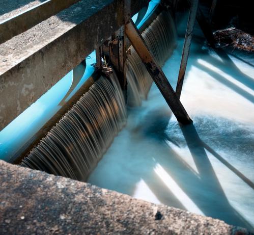 Auf dem Foto ist eine kleine Wasserwehranlage zu sehen. Das Wasser fließt gleichmäßig über eine Kante und bildet einen sanften, seidigen Effekt, der  durch eine Langzeitbelichtung erzielt wurde. Metallische Trägerkonstruktionen stützen die Struktur, und das Licht wirft deutliche Schatten auf die Wasseroberfläche. Die Szene wirkt ruhig und technisch zugleich.