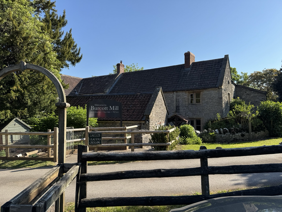 The view of an old two storey stone building that is the Burcott mill. The mill is long and has a peeked roof with two chimney stacks. The mill is surrounded by mature trees in full leaf.