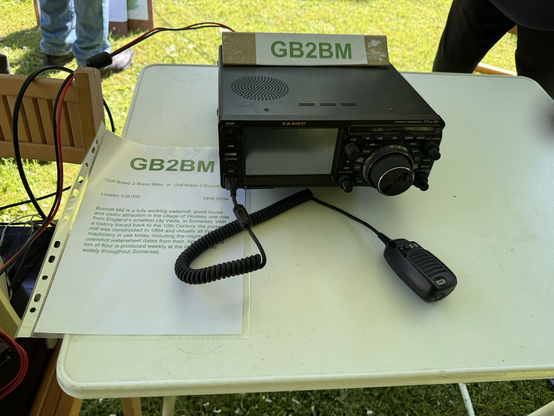 A black Yeasu FTdx10 radio sits on a white table. Balanced on top of the radio is the station call sign GB2BM. On the left hand side of the table is an information sheet about Burcott flour mill