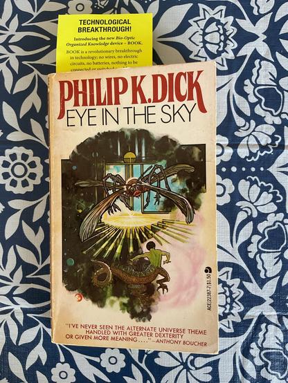 A photo of a paperback edition of the novel Eye in the Sky by Philip K Dick set against a blue and white floral backdrop.
