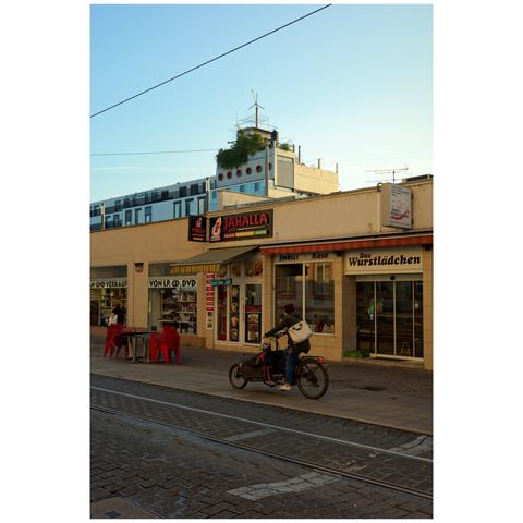 A person with a cargo bike is driving in front of a few stores in a low building. In the background, a larger building with a wind wheel on top.