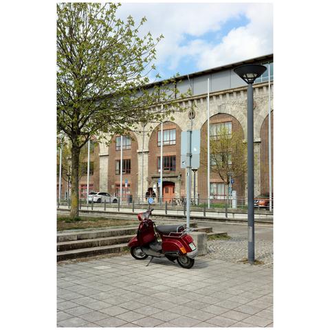 An older, dark red, classical scooter is parked in the corner of a square. In the background is a historic building, and on the left is a tree with fresh leaves.