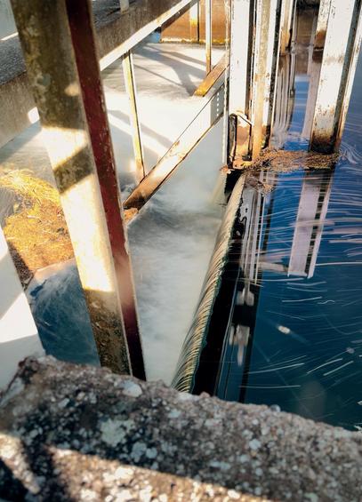 Auf dem Foto ist eine Wasserwehranlage zu sehen. Das Wasser fließt durch eine Konstruktion aus Metall- und Betonstrukturen, wobei es über eine Kante strömt und dabei eine glatte, weiße seidige Strömung erzeugt. Die metallenen Stützen zeigen Rostspuren, was auf eine längere Nutzung und Feuchtigkeitseinwirkung hinweist. Rechts im Bild ist ruhigeres Wasser mit Spiegelungen der umgebenden Strukturen zu erkennen.