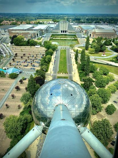 This image depicts a unique aerial view of the Atomium in Brussels, Belgium. The Atomium is a landmark building constructed for the 1958 Brussels World’s Fair (Expo 58). It’s made up of several spheres connected to form the shape of a unit cell of an iron crystal magnified billions of times. The structures in the background include part of the Brussels Expo exhibition halls, characterized by their large, expansive flat roofs and surrounded by parking areas and green spaces. The image captures the distinctive architecture and symmetry of the site, as well as the urban layout surrounding it.