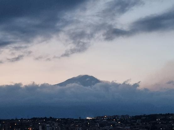 Etna quasi totalmente avvolta tra le nuvole, mentre il sole è già tramontato e le luci iniziano ad accendersi nella città ai suoi piedi