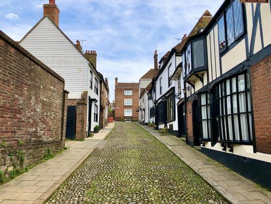 This image shows a charming cobblestone street lined with historic, timber-framed buildings. The buildings exhibit a Tudor architectural style, characterized by black and white exteriors, steeply pitched roofs, and prominent cross-beams. There's also a brick building with what appears to be an alley alongside it. The street is empty, giving the scene a quaint and serene look, typical of a traditional English village. The greenery and bright sky add a fresh atmosphere to the scene. Signs on the buildings suggest some are commercial, perhaps shops or restaurants, which contribute to the local economy and tourism.