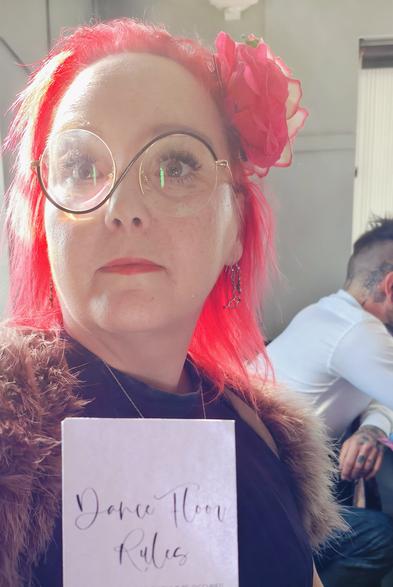 MrsVark, a woman with vibrant electric red hair and round glasses, sporting a red flower tucked behind her ear. She's wearing a fluffy jacket and holding a card that appears to read 'Dance Hoor Rules' (Dance Floor Rules in a cursive script) whilst giving the camera a rather unimpressed look.