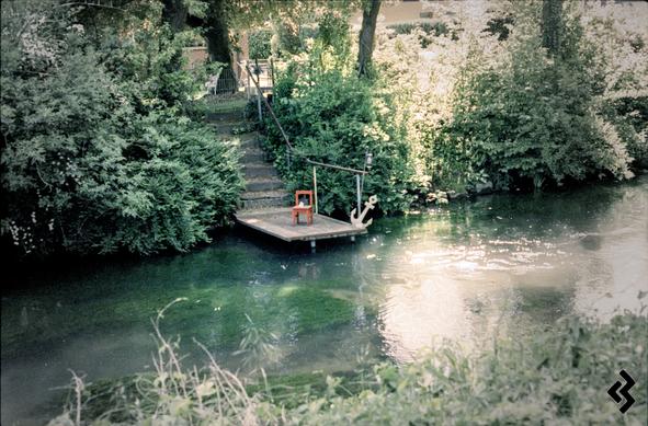 A small wooden dock with a tiny red chair and a wooden anchor replica sits at the edge of a clear, green-tinged river surrounded by dense foliage and trees; stone steps lead up from the dock into a garden area.