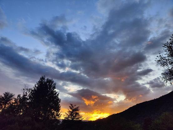 Soleil couchant se reflétant dans les nuages : offrant un dégradé, du jaune intense jusqu'au gris, alors que les nuages se déploient dans un ciel bleu en arrière-plan. Des arbres à contre-jour déjà sombres au premier plan.