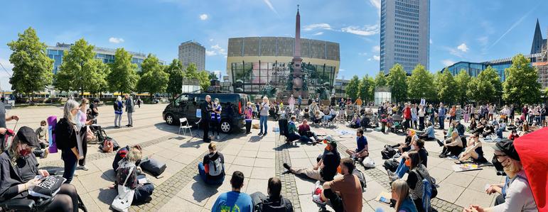 Das Bild zeigt die LiegendDemo 2025 auf dem Augustusplatz in Leipzig. Bei sonnigem Wetter liegen und sitzen viele Menschen entspannt auf dem Boden, einige mit Schildern oder Transparenten. In der Mitte steht ein schwarzer Transporter mit Lautsprecheranlage; davor spricht eine Person. Im Hintergrund sind der Mendebrunnen, das Opernhaus und der Uni-Riese zu sehen. Die Stimmung wirkt ruhig und solidarisch.