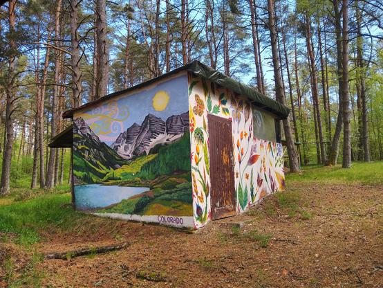 Budynek w lesie, pokryty muralami.

Na ścianie lewej, pejzaż z podpisem "Colorado", przedstawiające jezioro pośród gór. Jezioro jest błękitnobiałe, otoczone zielono-żółtymi pagórkami. Po prawej stronie ciemnozielony las. Z tyłu góry — po lewej stronie pokryte zielenią, po prawej skaliste. U góry błękitne niebo, z palącą żółtą kulą słońca po prawej stronie, i żółto-białymi spiralnymi wzorkami po stronie lewej.

Ściana frontowa ozdobiona jest różnobarwnymi rysunkami roślin, kwiatów i szmeterlingów na kremowobiałym tle. Znajdziemy odcienie czerwieni, pomarańczu, żółci, zieleni i błękitu, fioletu i koloru liliowego. Po lewej stronie tej ściany oszrupane drzwi, w prawym górnym rogu okno z pleksy.

Dookoła budowli ziemia pokryta brązowym igliwiem oraz zieloną trawą. Z tyłu las, pomiędzy pniami sosen widać zieleń liściastych krzewów, a u góry błękit nieba.