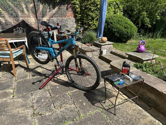 A n awesome blue mountain bike is mounted on a repair stand in a sunny garden, with various tools and maintenance supplies on a small table beside it. Outdoor furniture and greenery are visible in the background.