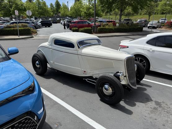 A classic white coupe car with a vintage design is parked between two modern vehicles in a parking lot. The car features a distinctive front grille and large, rounded tires. Lush greenery is visible in the background.