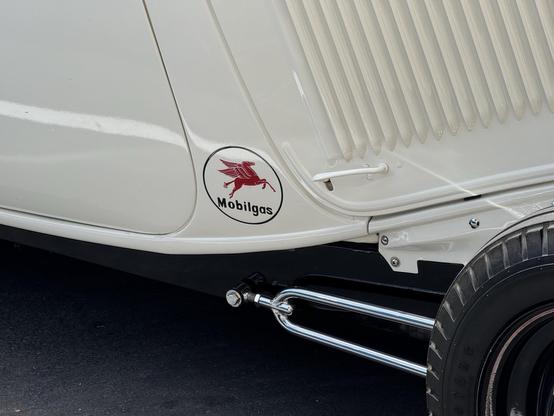 A vintage car detail featuring a Mobilgas logo with a red flying horse, positioned on a white car body. The image shows chrome exhaust pipes and a section of the car's design.