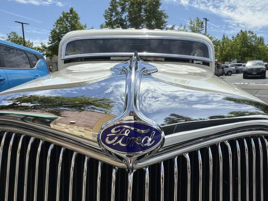 Image features the front view of a vintage Ford car, showcasing a shiny chrome grille and hood ornament. The background includes trees and parked vehicles under a blue sky with clouds.