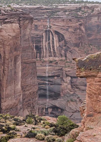 A color portrait photo of a thin waterfall falling from what is normally a dry wash probably 500-600 feet straight down a red-brown sandstone cliff face. There are an array of vertical black mineral stripes on the rock face just below the top of the falls. There are vertical cliffs on the left and a cliff top juts out on the right. In the lower foreground are a few scrubby plants on the cliff edge.