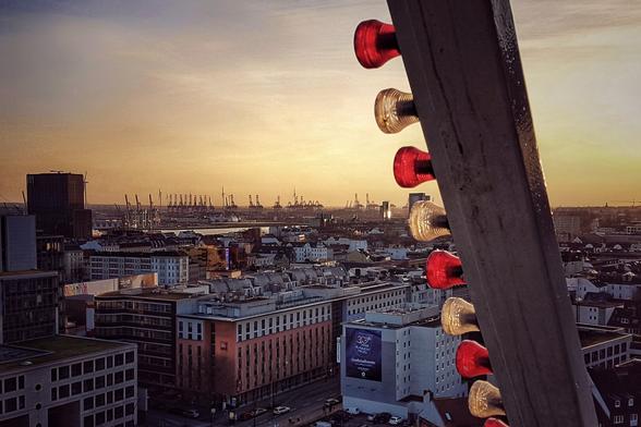 A high-angle view of a cityscape at sunset, taken from a Ferris wheel. The sky is a gradient of orange and yellow. City buildings and the dockland cranes are visible in the distance. The side of the Ferris wheel with illuminated red and clear lights is in the foreground on the right.