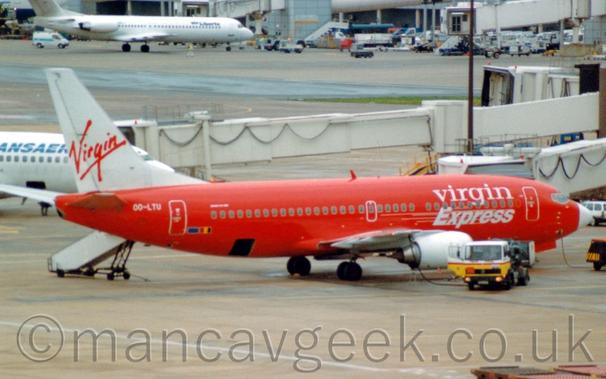 Side view of a red and white, twin engined jet airliner parked facing to the right.
The plane has a red body, with white "Virgin Express" titles on the forward fuselage, and a white nose cone.
The tail is white, with red, seemingly hand-written "Virgin" titles.
A yellow and white fuelling truck is parked next to the white engine under the right wing, with a fuel hose running into one of the wings internal fuel tanks.
Grey concrete apron fills the foreground, long, cream-coloured airbridges stretching across the frame in the background from the right, one attached to this plane, another to a white plane behind it on the left.
More concrete apron leads off to a grey terminal building in the distance at the top of the frame, with a white jet airliner parked next to it.