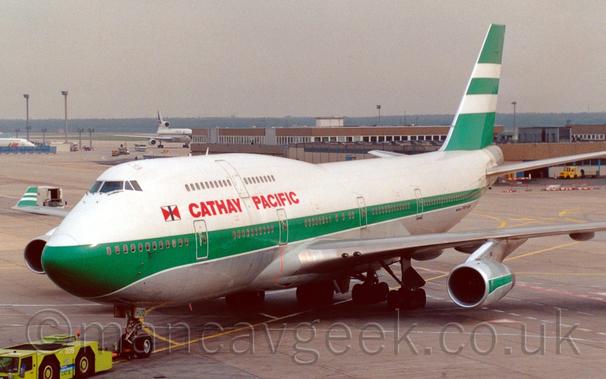 Side view of a very large, white and green, 4 engined jet airliner facing to the left but being pushed back to the right by a lime green tug attached to the main wheel by a long orange pole.
The plane is mostly white, with a thick green stripe running along the body, covering the passenger windows on the lower deck, and a grey belly, with white piping.
The tail is mostly green, with 2 thick white stripes in the middle.
The engine pods under the wings are grey, with a thick green stripe.
There are red "Cathay Pacific" titles on the upper forward fuselage, between the upper and lower deck windows.
A brown building stretches across the frame from the right in the background, with a white, 3 engined jet airliner just taxiing behind it.
Trees line the horizon, under grim grey skies.