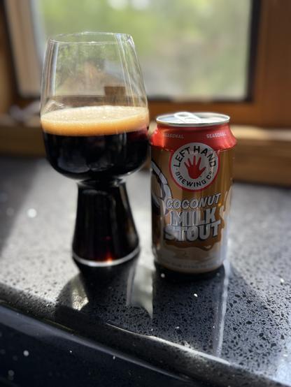 A tall stout glass is bathed in sunlight next to a 12 ounce can with a red and white label and the red LeftHand logo top center