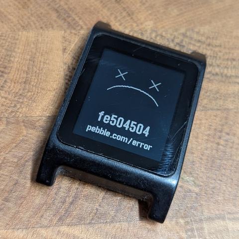 An old Pebble 2 watch without its straps and with a sad face and a fe504504 error message on the screen.