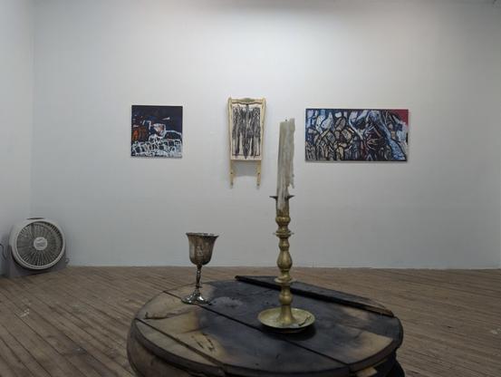 A candle on a blackened table in the foreground. Three darkly abstract paintings in the background