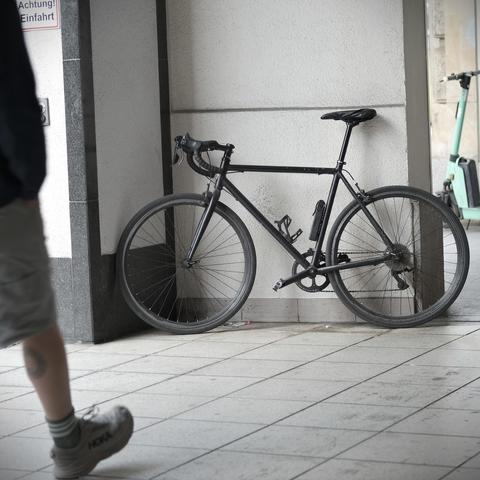 Ein schwarzes Rennrad in all-black, mit Lackschäden lehnt an einem Arkadenpfeiler. Ein Mensch läuft ins Bild.