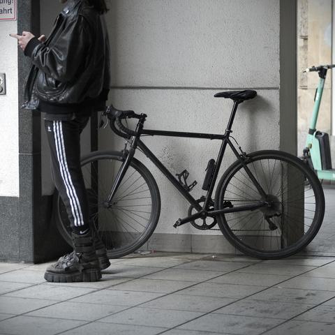 Das Paar aus Fahrrad und Fahrer. Er steht am Smartphone, trägt eine schwarze Lederjacke, eine schwarze Sporthose mit drei weißen Streifen, schwarze Socken, massive schwarze Stiefel und seine Fingernägel sind schwarz lackiert.