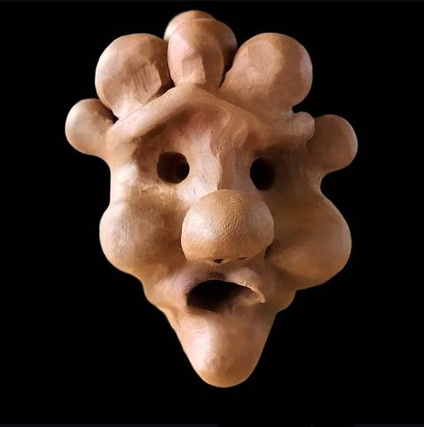This image shows a light beige clay or ceramic sculpture with an abstract face-like appearance against a black background. The sculpture has a rounded, bulbous form with several organic protrusions at the top that resemble hair or crown-like elements. It features two circular holes that appear as eyes, a prominent rounded bulge in the center that resembles a nose, and an opening at the bottom that suggests a mouth or jaw. The texture appears smooth with subtle contours that create a somewhat anthropomorphic or mask-like quality. The lighting highlights the sculpture's dimensional features and natural clay coloration.