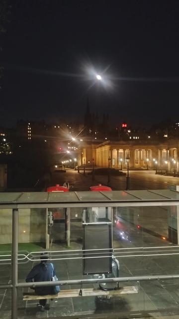 View from upper deck of a double decker bus, looking over a bus shelter to the illuminated National Gallery of Scotland and the Old Town, with a bright Moon above