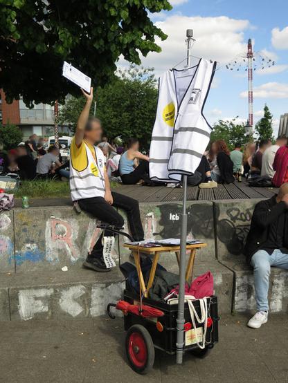 Mann mit weißer Warnweste mit #HamburgWerbefrei Logo sitzt auf einer Mauer im Park fiction. ER hält eine Unterschriftliste hoch.Vor ihm ein kleiner Wagen (Fahrradanhänger) mit weißer Warnweste mit Hamburg-Werbefrei Logo als Fahne.