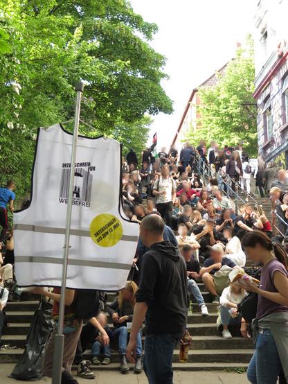 Eine weiße Warnweste mit #HamburgWerbefrei Logo hängt wie eine Fahne an einer Art Mast vor einer Menschenmenge. Die Menschen sitzen sehr nahe beieinander auf der Balduin-Treppe.