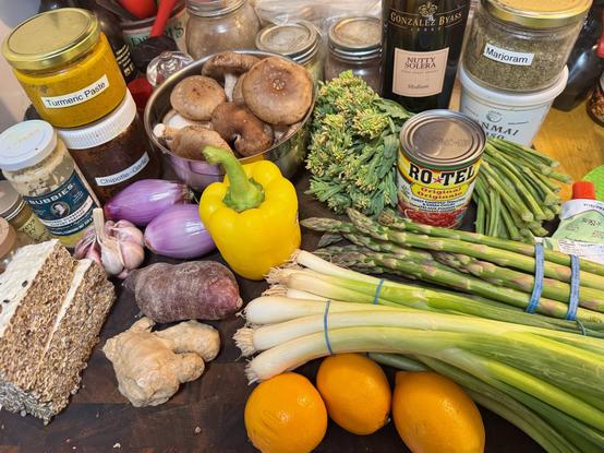 A cutting board on which are placed two blocks of homemade tempeh, a head of red garlic, two scallions, a jar of Bubbies horseradish, a jar of homemade turmeric paste, a jar of homemade chipotle-garlic paste, a bowl of large shiitake mushrooms, a yellow bell pepper, a purple-fleshed potato, a second of ginger root, three Meyer lemons, three bunches thick scallows a bunch of asparagus, a can of Ro•Tel Original, a botlle of Nutty Solera sherry, a jar labeled marjjoram, a small container of genmai miso, a rolled-up tube of tomato paste, and a bunch of kale sprouts.