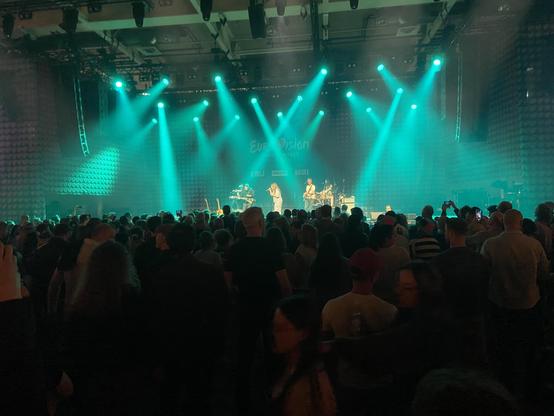 Anna Rossinelli und Band singt zum Auftakt des #ESC2025 in der Messehalle in Basel unter einem türkis grünen Lichtermeer im Scheinwerfer Licht