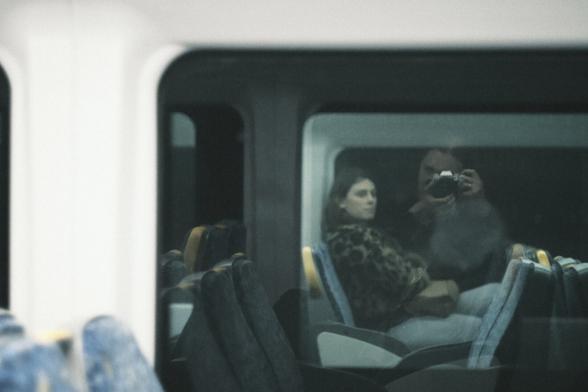 A photo from one train passing another late at night. The shot is in washed out colour and shows the only woman visible in the carriage, glancing across into the other train. Reflected in the window is the photographer, a woman with red hair looking through the viewfinder of the camera.
