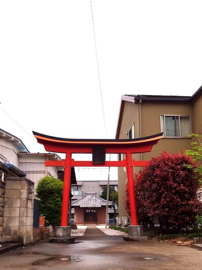A red wooden gate standing in between residential housing. The shrine is in the background, some 80m or 240ft back. The sky is gray.