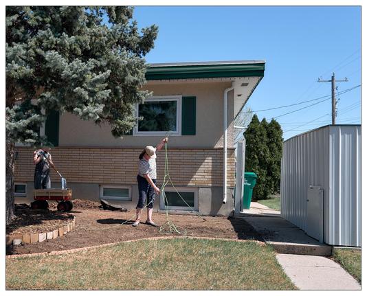 Two individuals engaged in yard maintenance. One person appears to be adjusting or installing a green electrical cord or maybe a hose.  The other person is near a small wagonin a driveway, shovelling dirt.

The setting includes a house with a visible window adorned with green shutters, and a neat yard bordered by bricks likely designating a garden or planting area. There's also a sidewalk running along the property edge, a metal storage shed, and a trash bin. The environment suggests a clear, sunny day with blue skies, which is ideal for outdoor chores.