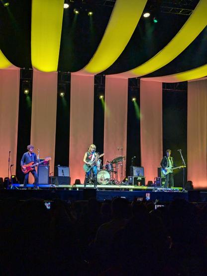 Una banda de rock está tocando en un escenario con una iluminación dramática. El escenario está decorado con cortinas verticales en blanco y amarillo pálido, creando un patrón de rayas. Tres miembros de la banda son visibles: un guitarrista a la izquierda, una cantante y guitarrista en el centro y un guitarrista a la derecha. Todos están tocando sus instrumentos. Detrás de ellos hay una batería y equipo de amplificación. Un público numeroso se encuentra sentado en la oscuridad frente al escenario.