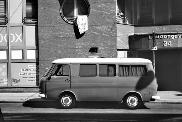 The black and white image shows a vintage van parked on the street in front of a modern building with a round window above, from which a blanket or quilt is hanging out to dry.