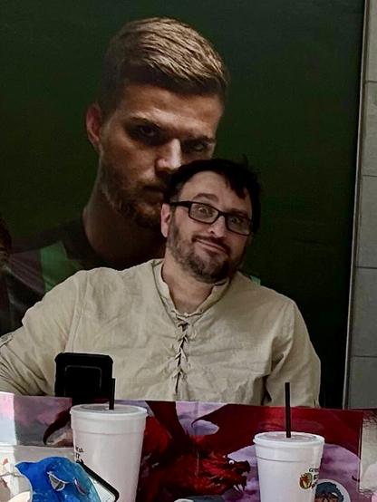 Me, a bearded guy wearing a tan colored medieval fantasy pirate shirt thing, sitting behind a Dungeons & Dragons Dungeon Master screen. Behind me, a mural of some dude is positioned just right that it looks like he is sniffing my hair.