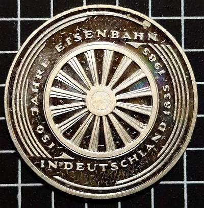 1985 5 Deutsche Mark Eisenbahn in Deutschland
Steam locomotive wheel. Inscription that celebrates the 150 years of the Germany railroad. The first railroad line was open on December 7th, 1835 between Nuremberg and Fürth

Script: Latin

Lettering: 150 JAHRE EISENBAHN IN DEUTSCHLAND 1835-1985

Engraver: Erich Ott