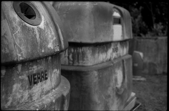 Black and white photography of glass and paper container. The glass container is in the foreground and the paper one is in the background and slightly out of focus. The are old and mossy.