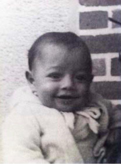 A face of a smiling baby boy. He is photographed against a brick wall background.