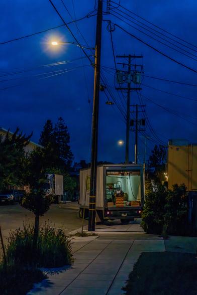 A night photograph of a truck unloading on an empty street. The dark blue sky is illuminated by a yellow streetlamp and the light blue artificial light inside the truck.