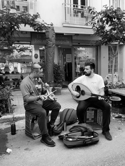 Black and white photograph. Two men sitting on upturned plastic crates on the asphalt during a street market, busking. One is playing a lyra, the other a laouto, and they are communicating with music and their gaze, in that wonderful way that musicians have. It’s this, as much as the traditional instruments, that made me photograph them.
They are on Kalidromiou in Exarcheia on a Saturday