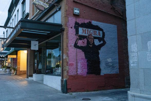 A photograph of a pink and white mural on a brick wall. A black outline with highlighted features of a woman dominates the center of the mural, and she appears to be carrying a large bundle labeled "MIGRA".