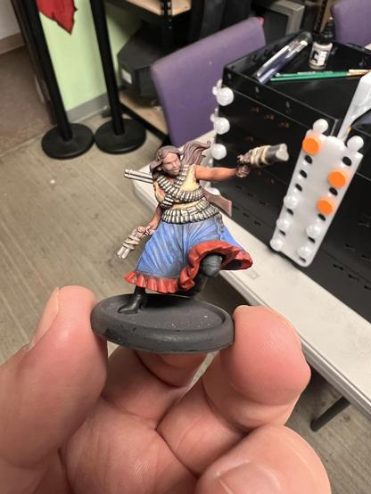 A painted miniature of a Monster Hunter from the game Malifaux. The mini features a heavier set Hispanic woman dual wielding triple barreled pistols as she jumps into battle with a rifle on her back.