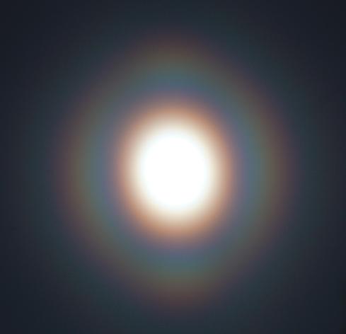 Um den überbelichteten hellen Mond zeigt sich ein rotbräunlicher Hof, umgeben von einem regenbogenfarbigen Ring. Der Ring hat hellere Knoten oben, unten, links und rechts.